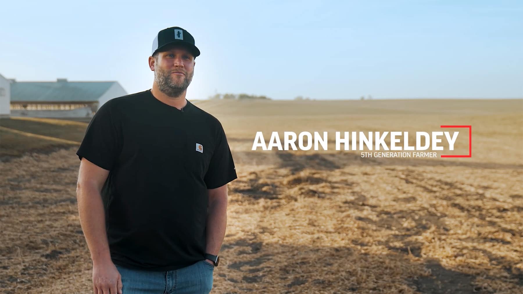 Farmer in t-shirt, jeans and hat stands in harvested field with pig barns behind him.